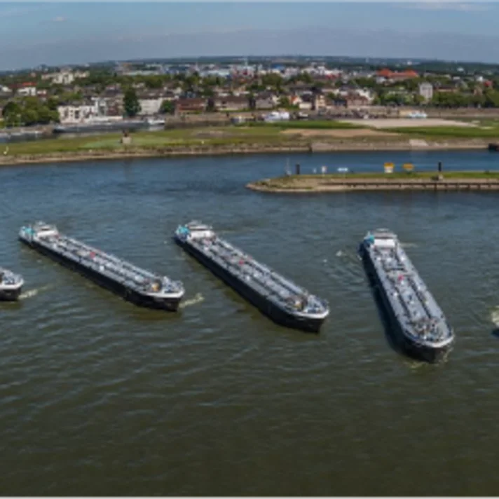 Mehrere Frachtschiffe liegen nebeneinander auf einem breiten Fluss, während eines den Verband verlässt. Im Hintergrund sind eine Brücke, grüne Uferbereiche und eine Stadtlandschaft mit Industriegebäuden zu sehen.