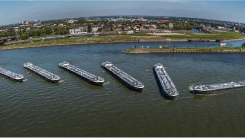 Mehrere Frachtschiffe liegen nebeneinander auf einem breiten Fluss, während eines den Verband verlässt. Im Hintergrund sind eine Brücke, grüne Uferbereiche und eine Stadtlandschaft mit Industriegebäuden zu sehen.
