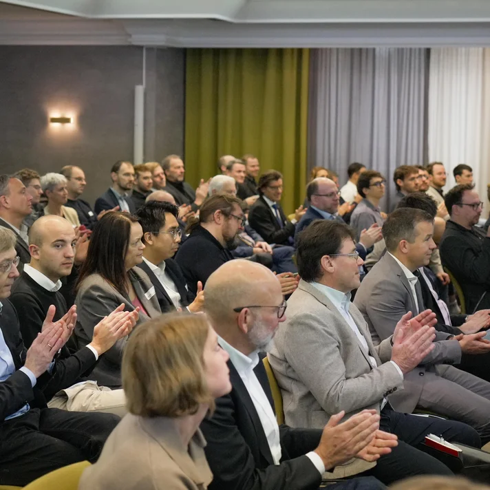 Mehrere Personen sitzen in Reihen in einem Konferenzraum und klatschen, einige tragen Anzug oder Business-Kleidung.