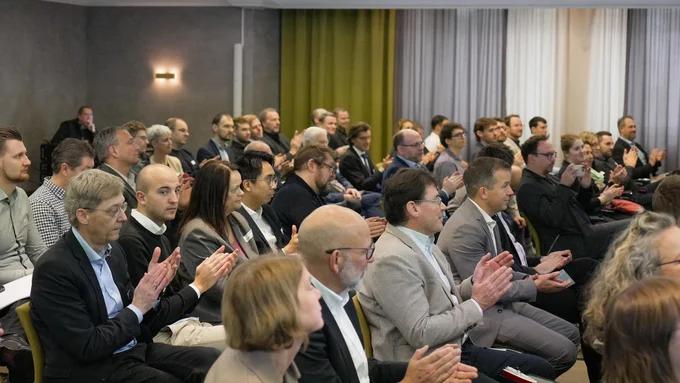 Mehrere Personen sitzen in Reihen in einem Konferenzraum und klatschen, einige tragen Anzug oder Business-Kleidung.