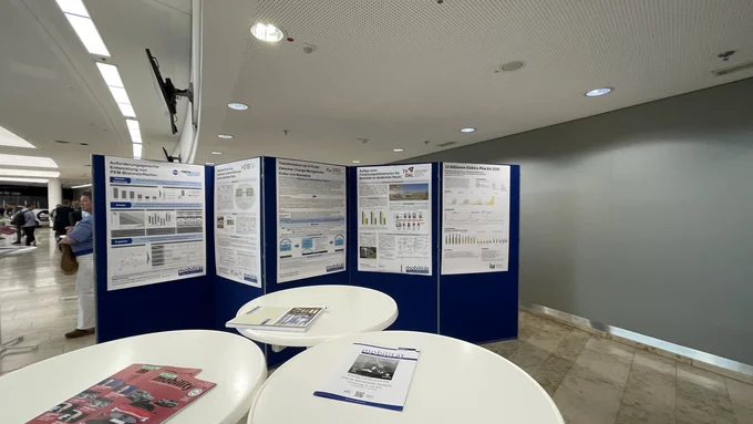 Drei weiße Stehtische hinter denen eine blaue Faltwand mit fünf Projektpostern steht.