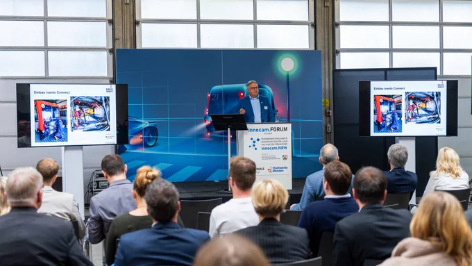 Publikum hört einem Vortrag beim innocam.FORUM zu, mit Präsentation und großem Hintergrundbild eines autonomen Busses.