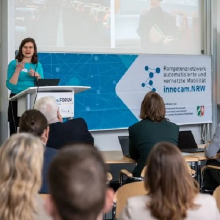 Eine Frau steht an einem Rednerpult mit einem Banner des Netzwerks "innocam.NRW" und spricht vor einem Publikum.