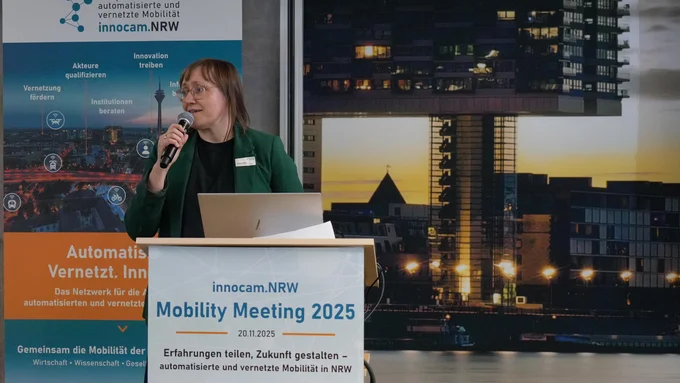 Frau mit Mikrofon steht an einem Rednerpult mit Schild 'innocam.NRW Mobility Meeting 2025' vor einem Banner des Kompetenznetzwerks automatisierte und vernetzte Mobilität und einem nächtlichen Stadtpanorama mit beleuchteten Gebäuden.