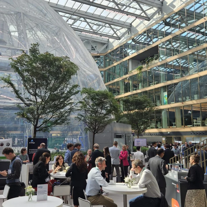 Sehr hoher Innenraum mit einer darin untergebrachten durchsichtigen Kuppel im Hintergrund, rund darum sieht man viele Personen, die an Stehtischen netzwerken.