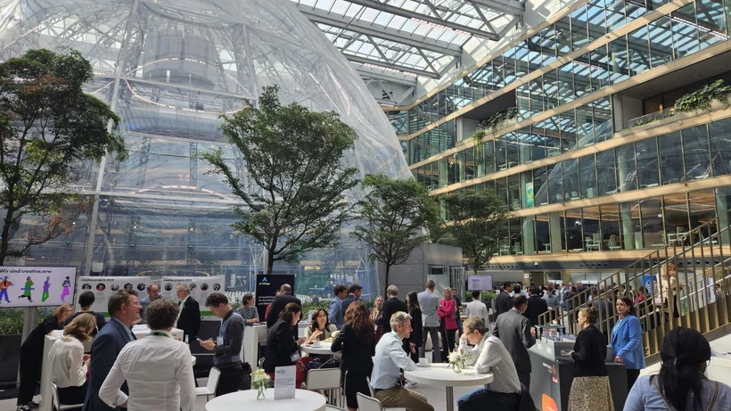 Sehr hoher Innenraum mit einer darin untergebrachten durchsichtigen Kuppel im Hintergrund, rund darum sieht man viele Personen, die an Stehtischen netzwerken.