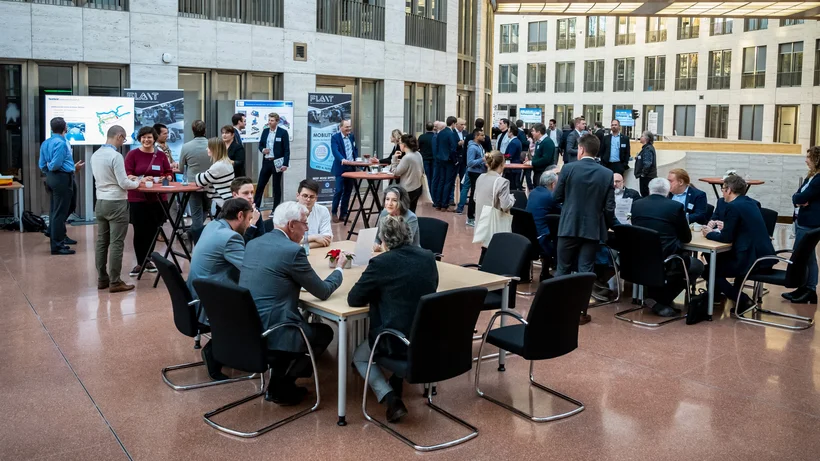 Teilnehmende eines Netzwerktreffens oder Fachkongresses im offenen Foyer eines modernen Gebäudes. Personen stehen an Stehtischen oder sitzen an Gruppentischen im Gespräch.