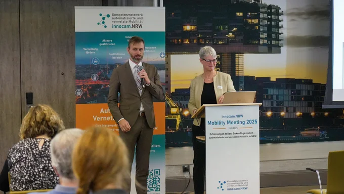 Zwei Personen stehen vor Publikum bei der innocam.NRW Mobility Meeting 2025, eine spricht am Mikrofon, die andere am Rednerpult mit Laptop.