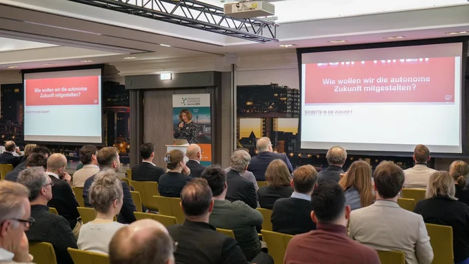 Konferenzraum mit Publikum, das einer Präsentation zum Thema autonome Zukunft folgt, sichtbar auf zwei Projektionsflächen mit dem Text 'Wie wollen wir die autonome Zukunft mitgestalten?'.