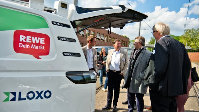 Gruppe von fünf Personen steht neben einem weißen Fahrzeug mit geöffnetem Heck, auf dem Fahrzeug sind die Logos 'REWE Dein Markt' und 'LOXO' sichtbar