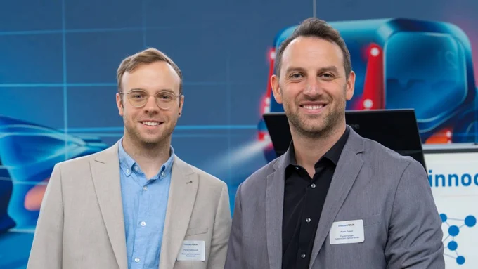 Zwei Männer in hellen Sakkos lächeln in die Kamera vor dem Hintergrund des „innocam.FORUM“.