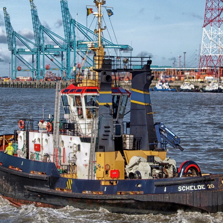 Schlepper mit dem Namen fährt auf einem Hafenbecken mit Kränen und Industrieanlagen im Hintergrund