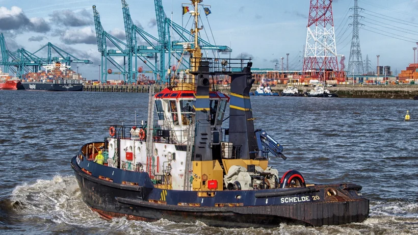 Schlepper mit dem Namen fährt auf einem Hafenbecken mit Kränen und Industrieanlagen im Hintergrund