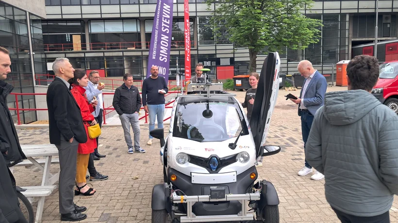 Kleines Elektroauto mit geöffnetem Türflügel umgeben von mehreren Personen auf gepflastertem Platz vor Bürogebäuden.