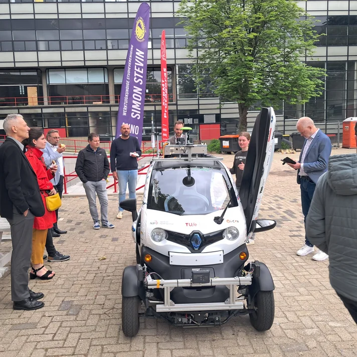Kleines Elektroauto mit geöffnetem Türflügel umgeben von mehreren Personen auf gepflastertem Platz vor Bürogebäuden.