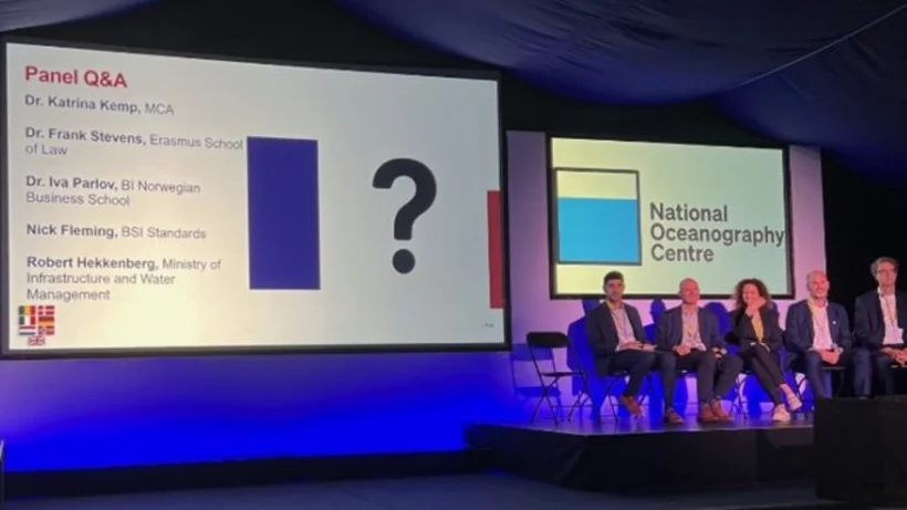 Eine pink und bläulich beleuchtete Bühne mit drei Leinwänden, die mittlere etwas größer als die zwei rechts und links, links steht eine Frau an einem Punkt und rechts sitzen fünf Personen auf Stühlen.