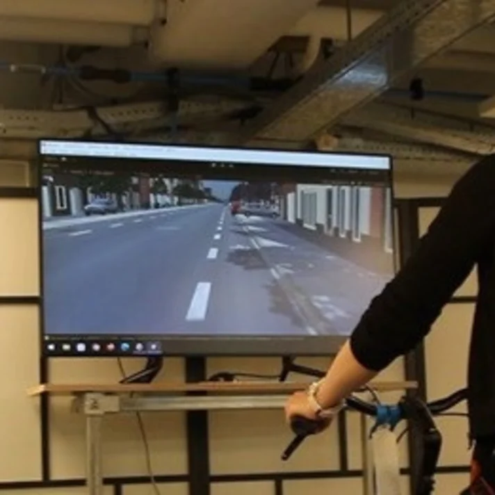 Person mit Helm auf einem Fahrradsimulator vor einem Bildschirm mit Straßenszene, zur Verkehrssimulation und Forschung.
