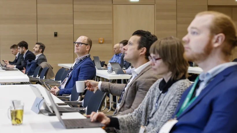 Personen sitzen in einem Konferenzraum an Tischen mit Laptops und Getränken und hören aufmerksam zu