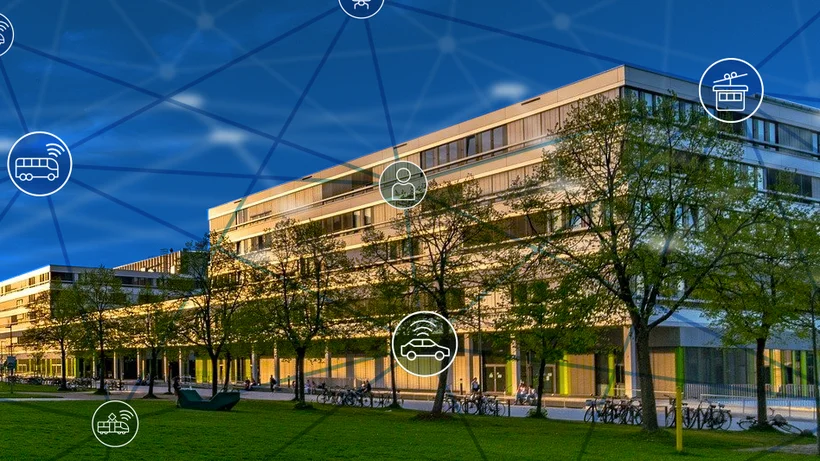 Modernes, mehrstöckiges Gebäude bei Sonnenuntergang, umgeben von Bäumen und Fahrrädern. Im Vordergrund ist eine Rasenfläche. Über das Bild sind transparente, vernetzte Symbole aus dem Bereich Mobilität (z. B. Auto, Bus, Bahn) gelegt, die eine digitale Vernetzung oder Smart-City-Technologie symbolisieren.