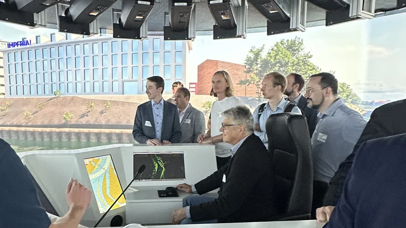 Zehn Personen stehn um den sitzenden Kapitän eines Schiffes. Das Schiff befindet sich augenscheinlich in einem Kanal. Im Hintergrund sind Bürogebäude zu sehen.