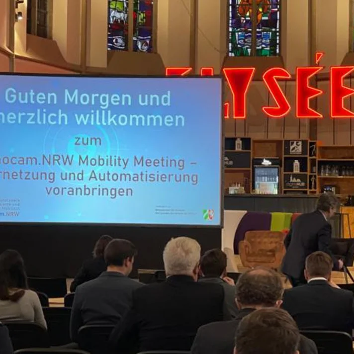 Ein kofnerenzsaal in einer ehemaligen Kirche, im hinteren Bereich eine leicht erhöhte Bühne, die eine Leinwand mit der Begrüßungsfolie der Veranstaltung zeigt. Daneben steht der Moderator der Veranstaltung Marcel Sonntag.