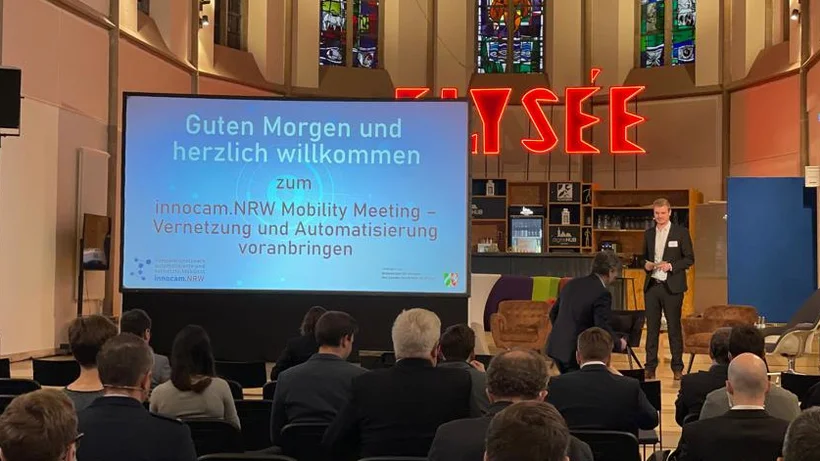 Ein kofnerenzsaal in einer ehemaligen Kirche, im hinteren Bereich eine leicht erhöhte Bühne, die eine Leinwand mit der Begrüßungsfolie der Veranstaltung zeigt. Daneben steht der Moderator der Veranstaltung Marcel Sonntag.