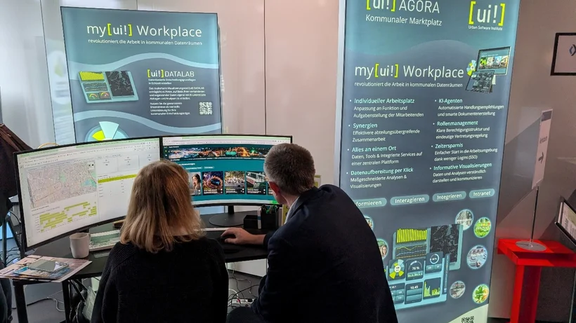 Zwei Personen sitzen vor drei Bildschirmen an einem Arbeitsplatz mit Bannern im Hintergrund, die die Software my[ui!] Workplace und AGORA kommunaler Marktplatz bewerben