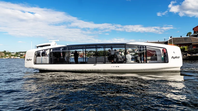 Modernes, langgestrecktes Passagierboot mit großen Panoramafenstern auf ruhigem Wasser vor Küstenlandschaft, seitlich sieht man die Aufschrift "hyke".
