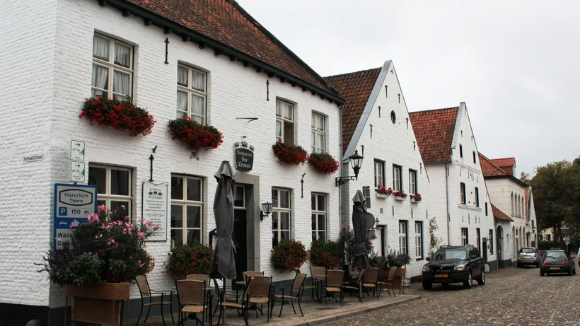 Straßenszene mit weiß verputzten Häusern, Blumenkästen an den Fenstern und Außengastronomie mit Tischen und Stühlen auf Kopfsteinpflaster
