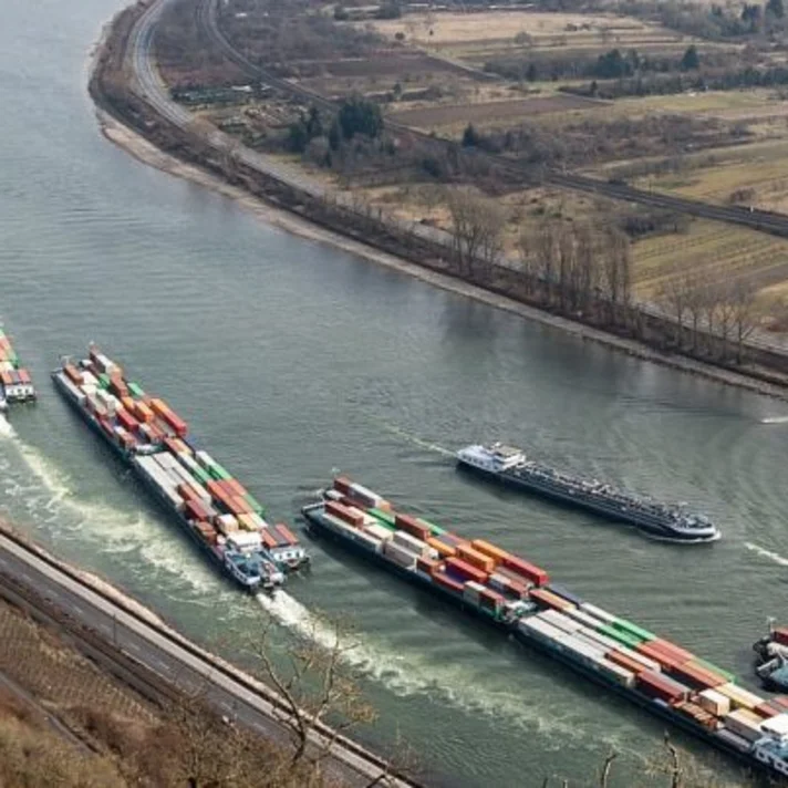 Mehrere beladene Containerschiffe fahren hintereinander auf einem kurvigen Fluss durch eine ländliche Gegend.