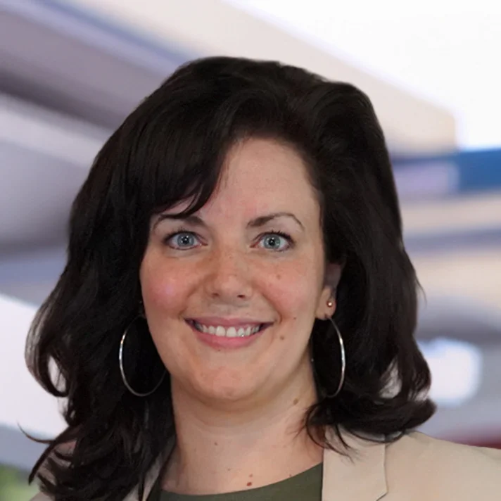 „Porträtfoto einer lächelnden Frau mit schulterlangem, dunklem, leicht gewelltem Haar. Sie trägt große runde Ohrringe, ein helles Sakko und ein dunkles Oberteil. Der Hintergrund ist unscharf und zeigt moderne Architektur in hellen Farbtönen.