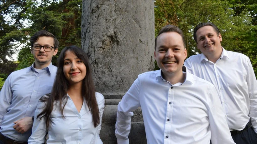 Vier junge Erwachsene posieren lächelnd vor einer Steinsäule im Park.