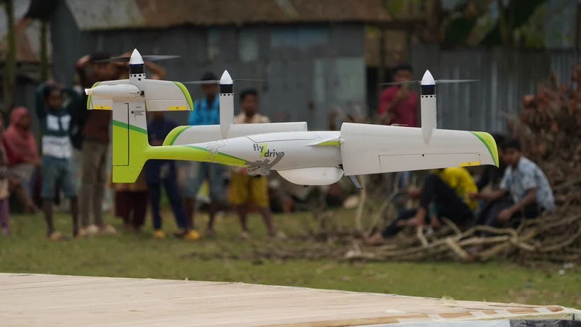 Eine weiß-grüne Lieferdrohne mit vier Rotoren schwebt knapp über einem Holzpodest. Im Hintergrund beobachten mehrere Menschen in ländlicher Umgebung das Geschehen.