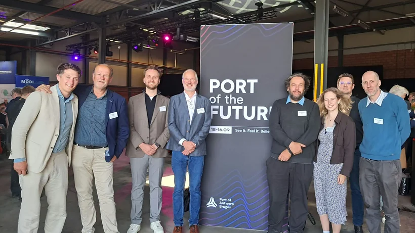 Sieben Personen stehen in einer Halle vor einem Banner mit der Aufschrift 'PORT of the FUTURE'