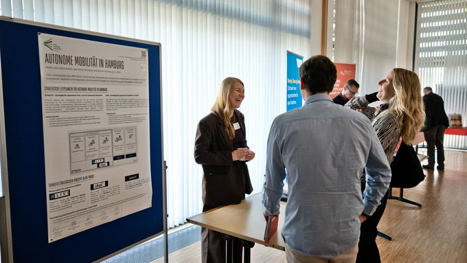 Personen stehen an einem Messestand mit einem großen Informationsplakat und einem Tisch in einem hellen Raum mit vertikalen Jalousien