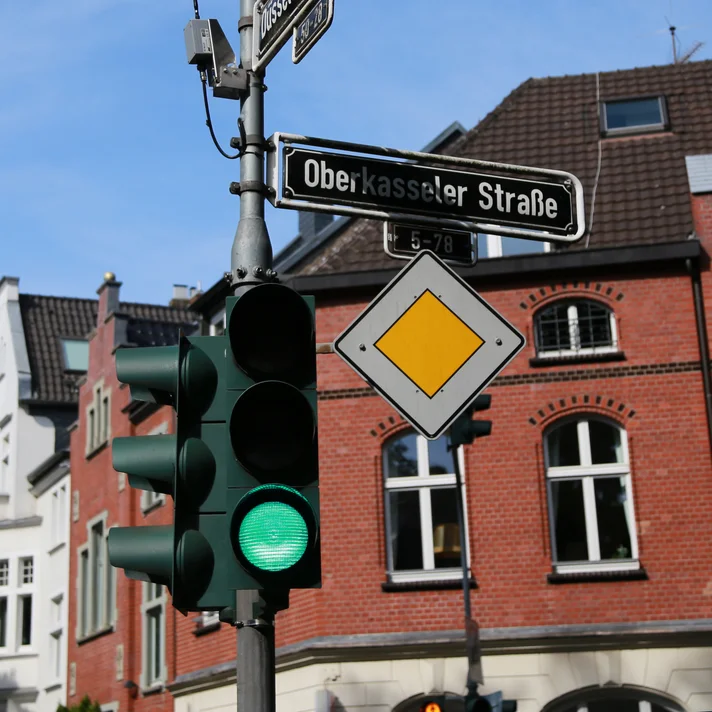 Eine Ampel an einer städtischen Kreuzung zeigt Grün. Darüber befinden sich Straßenschilder mit den Namen „Düsseldorfer Straße“ und „Oberkasseler Straße“. Ein gelbes Vorfahrtsschild ist ebenfalls sichtbar. Im Hintergrund stehen mehrstöckige Wohngebäude aus Backstein und Putz unter einem blauen Himmel.
