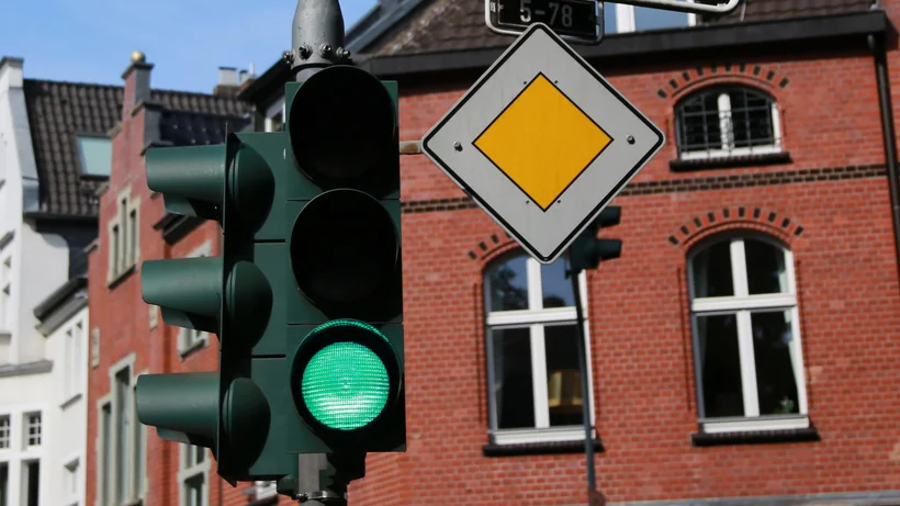 Eine Ampel an einer städtischen Kreuzung zeigt Grün. Darüber befinden sich Straßenschilder mit den Namen „Düsseldorfer Straße“ und „Oberkasseler Straße“. Ein gelbes Vorfahrtsschild ist ebenfalls sichtbar. Im Hintergrund stehen mehrstöckige Wohngebäude aus Backstein und Putz unter einem blauen Himmel.