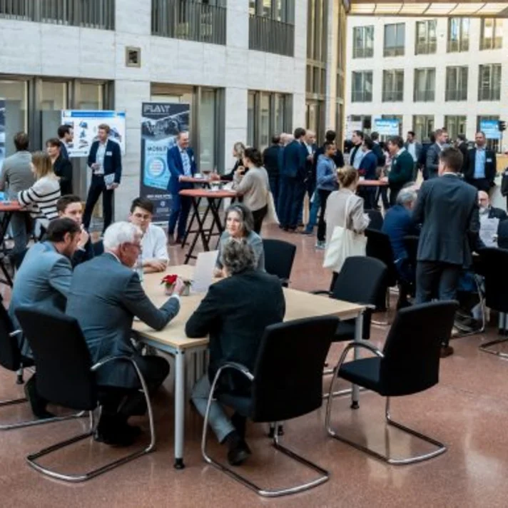 Menschen bei einer Netzwerkveranstaltung in einer offenen Halle im Gespräch und an Tischen.
