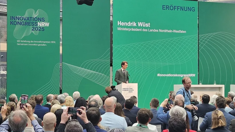 Ein Mann steht erhöht an einem Rednerpult, hinter ihm hohe, gräne Stehwände mit dem logo des Innovationskongress NRW 2025 und dem Namen Hendrik Wüst, Ministerpräsident des Landes Nordrhein-Westfalen, im Vordergrund eine Menschenmenge die ihm sitzend zuhört.