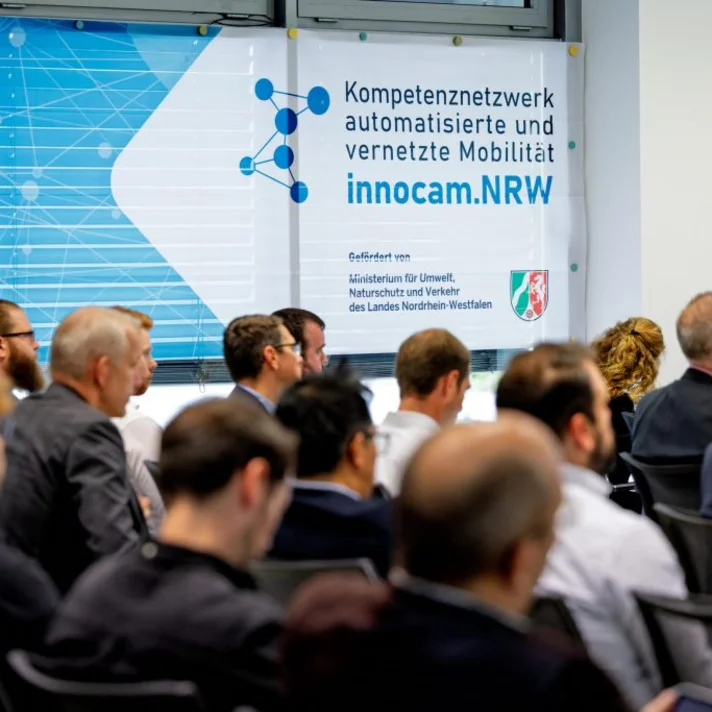 Mehrere Personen sitzen in einem Konferenzraum vor einer Wand mit einem Plakat, das das Kompetenznetzwerk 'innocam.NRW' für automatisierte und vernetzte Mobilität zeigt.