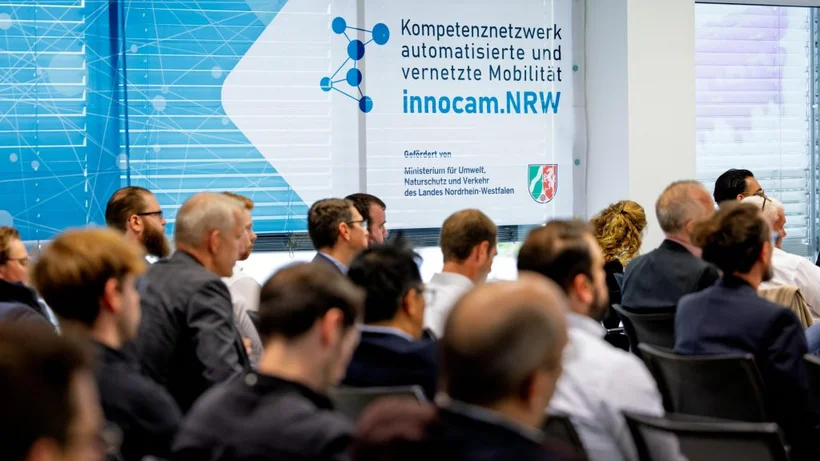 Mehrere Personen sitzen in einem Konferenzraum vor einer Wand mit einem Plakat, das das Kompetenznetzwerk 'innocam.NRW' für automatisierte und vernetzte Mobilität zeigt.