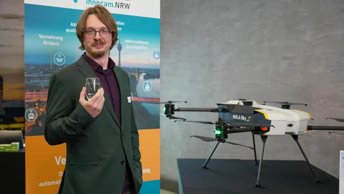 Mann in dunklem Sakko hält ein kleines Gerät, neben ihm steht eine Drohne mit der Aufschrift HHLA Sky auf einem schwarzen Podest, im Hintergrund ein Banner von innocam.NRW.