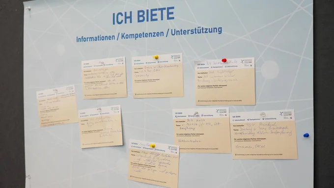 Tafel mit dem Titel 'ICH BIETE' und mehreren beige Zetteln, die Informationen, Kompetenzen und Unterstützung beschreiben, befestigt mit farbigen Pinnnadeln.