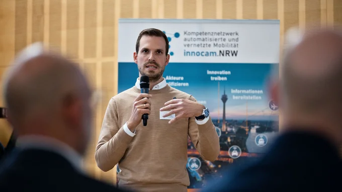 Mann mit Mikrofon vor Banner mit Text 'Kompetenznetzwerk automatisierte und vernetzte Mobilität innocam.NRW' spricht vor Publikum