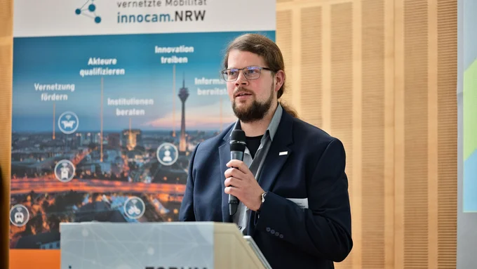 Mann mit Bart und dunklem Anzug hält Mikrofon vor einem Rednerpult mit Aufschrift 'innocam.FORUM' vor einer Wand mit Poster zum Kompetenznetzwerk automatisierte und vernetzte Mobilität innocam.NRW