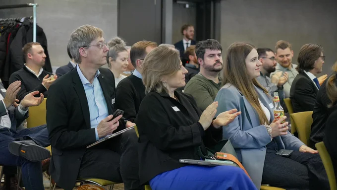 Mehrere Personen sitzen in einem Raum bei der innocam.NRW Mobility Meeting Veranstaltung, einige klatschen, eine Frau hält eine Flasche, andere haben Notizblöcke oder Smartphones in der Hand.