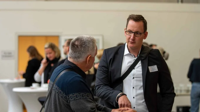 Zwei Männer sprechen am Stehtisch bei einer Konferenz, weitere Personen im Hintergrund.