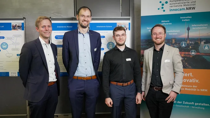 Vier Männer in Business-Kleidung stehen vor Informationsplakaten und einem Roll-up mit dem Logo und Text von innocam.NRW Mobility Meeting.