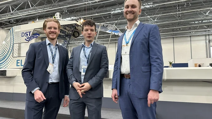 Drei Männer in Anzügen stehen in einer Messehalle und repräsentieren innocam.NRW auf der XPONENTIAL 2025.