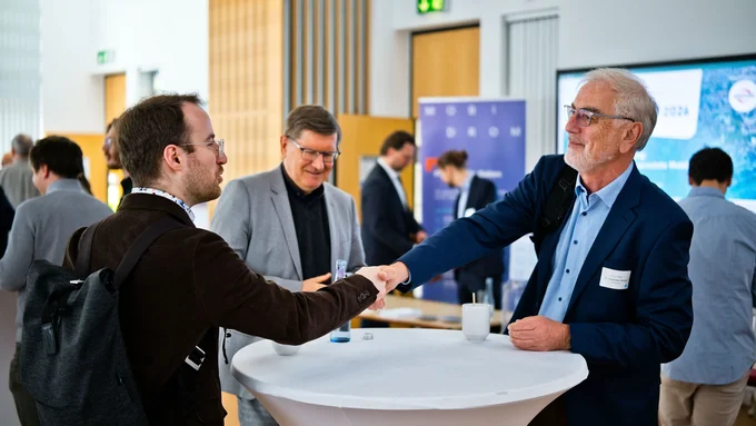 Zwei Männer geben sich an einem Stehtisch die Hand, im Hintergrund weitere Personen in einem hellen Raum mit Holzpaneelen und Informationsständen